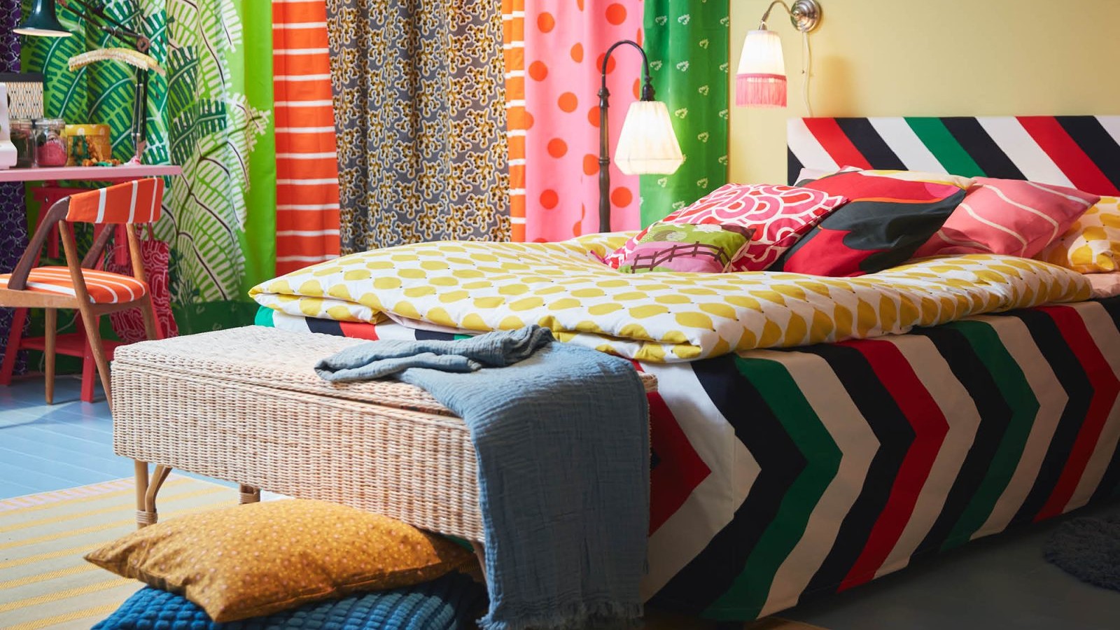 A vibrantly decorated bedroom features a bed with a colorful chevron duvet cover and a yellow patterned duvet, surrounded by numerous patterned textiles in bright colors.