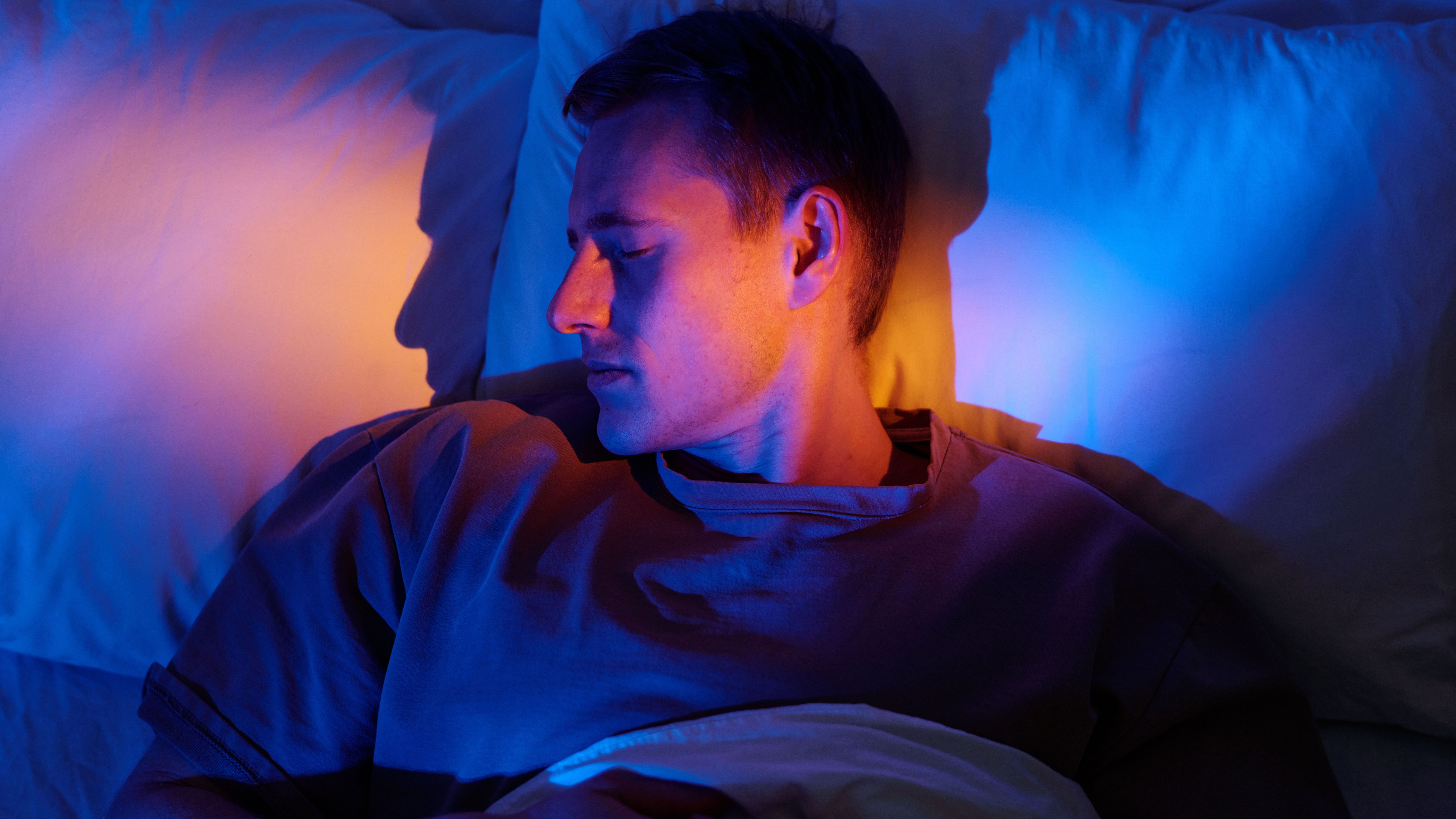 Man sleeping on bed with head resting on pillow, eyes closed and face illuminated by blue and orange light.