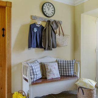 A pale yellow-painted hallway with a white bench with checked and floral cushions on top