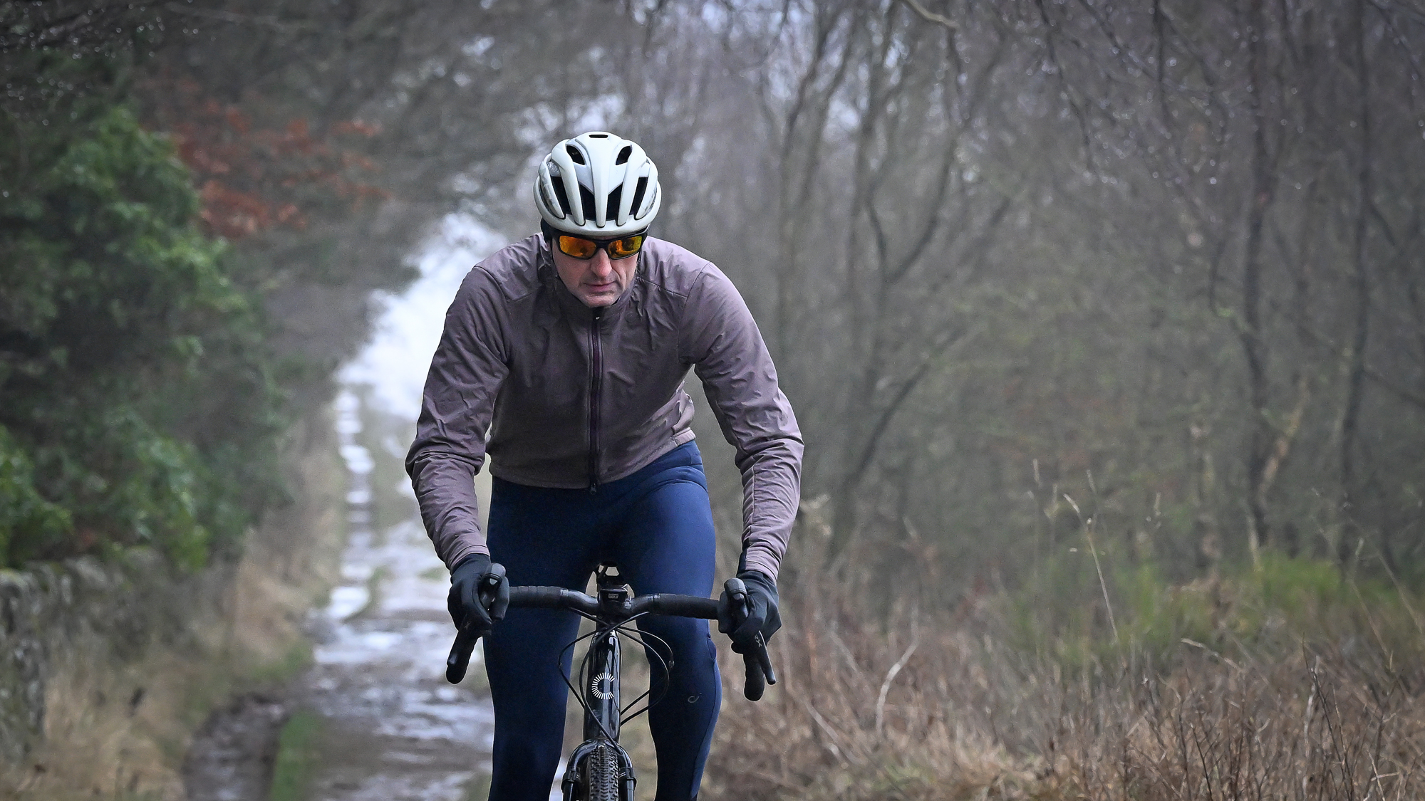 Man wearing a purple jacket, navy tights and a pale helmet riding a black gravel bike towards the camera on a muddy track
