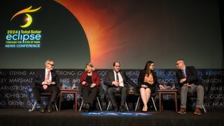 people wearing suits on a stage laugh as they hold paper eclipse glasses to their faces