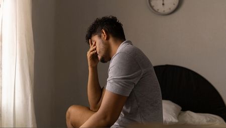 A man wearing a grey tshirt sitting on bed of bed in beige room with his head in his hands as he struggles with anxiety and depression and sleep.