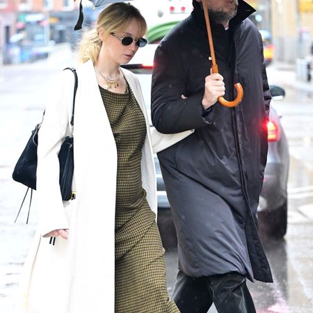 Jennifer Lawrence wears a oval sunglasses, black shoulder bag, white long coat, and green checkered maxi dress