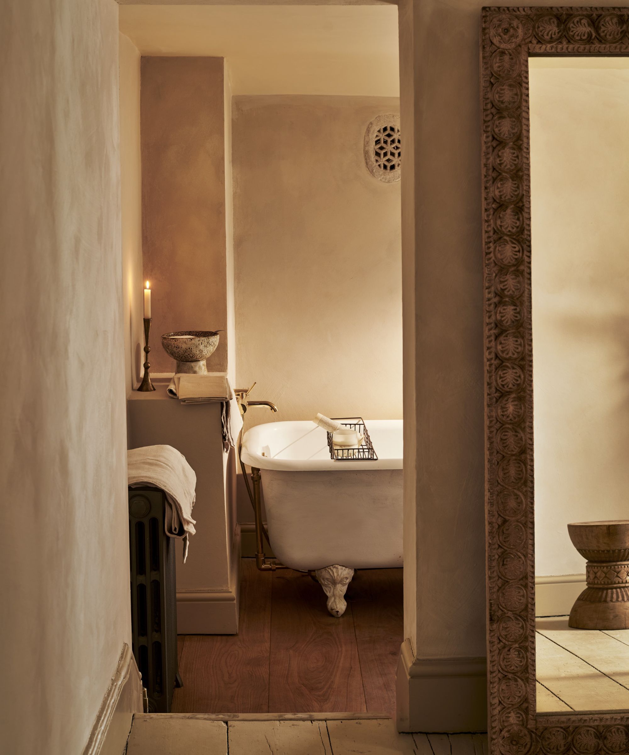 A softly lit bathroom with a freestanding roll-top bath, textured plaster walls, warm brass fittings and timber flooring.