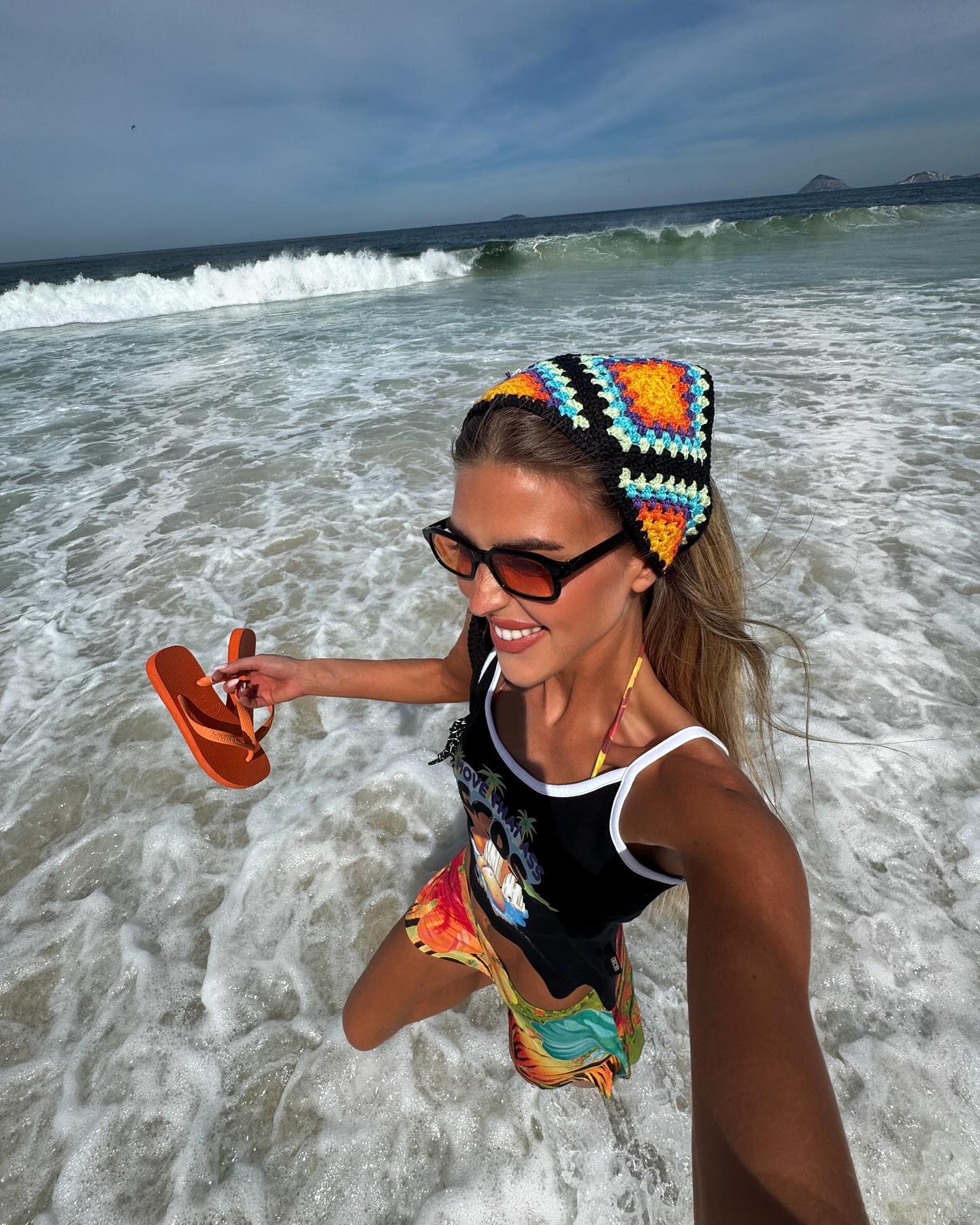 Influencer Emili wears a crochet headband, orange lensed glasses, a printed tank and tropical print skirt and is carrying orange Havaiana flip flops