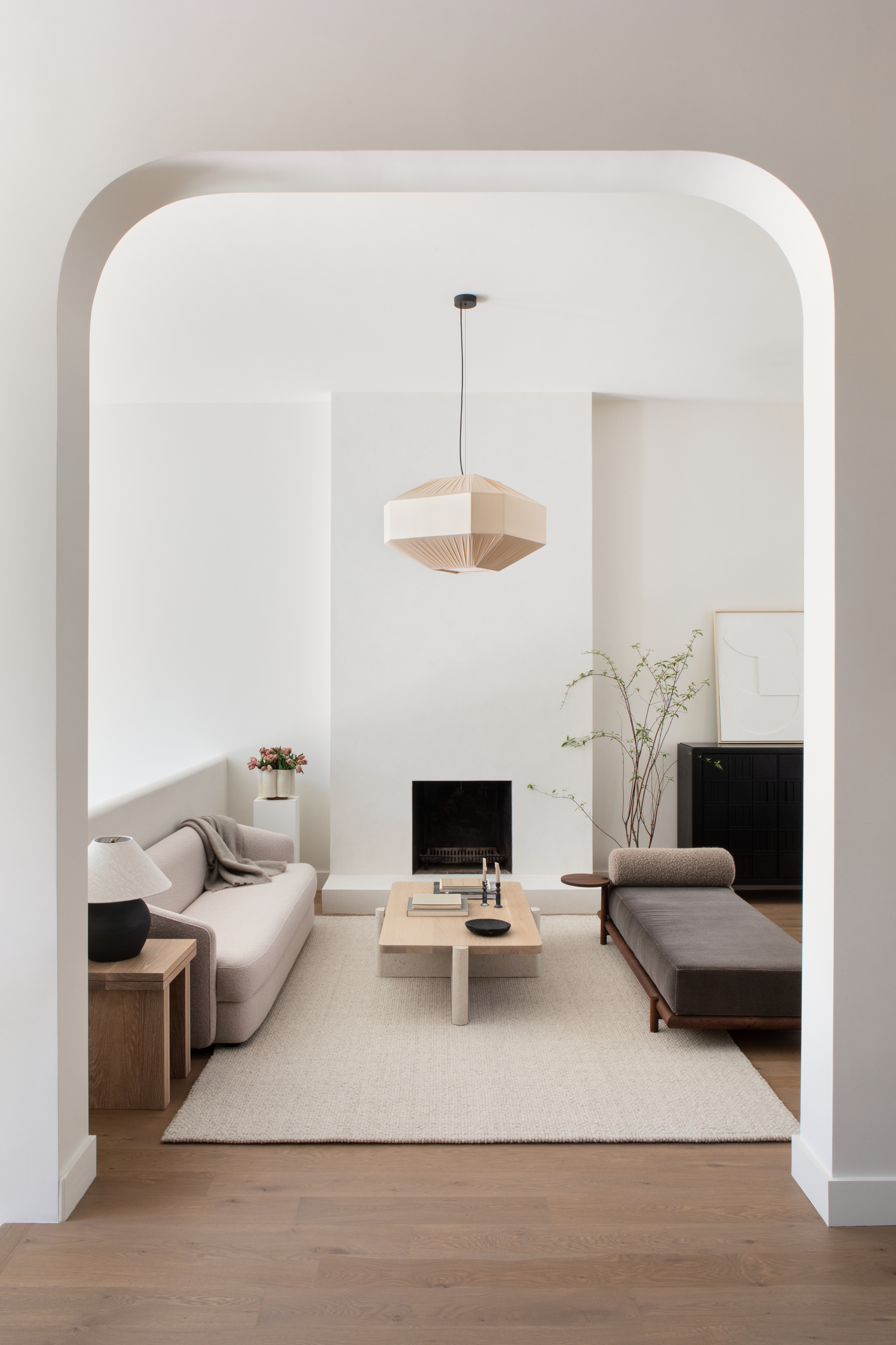 neutral living room with white walls, timber flooring, beige rug with timber coffee table, bench seat, beige sofa with throw, timber side table with lamp and white pedestal with flowers in vase, and string pendant light hanging from ceiling, and simple fireplace