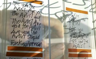 Two sheets of paper with black scribbled words hang on the wall, with footage of the artist creating them played over the top