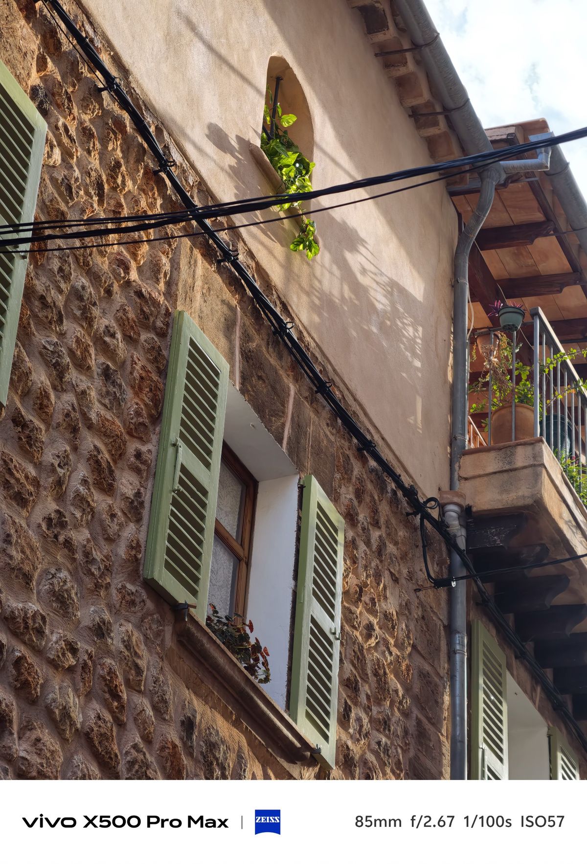 Upward view of a rustic stone house with pale green shutters, balconies, plants and overhead cables.