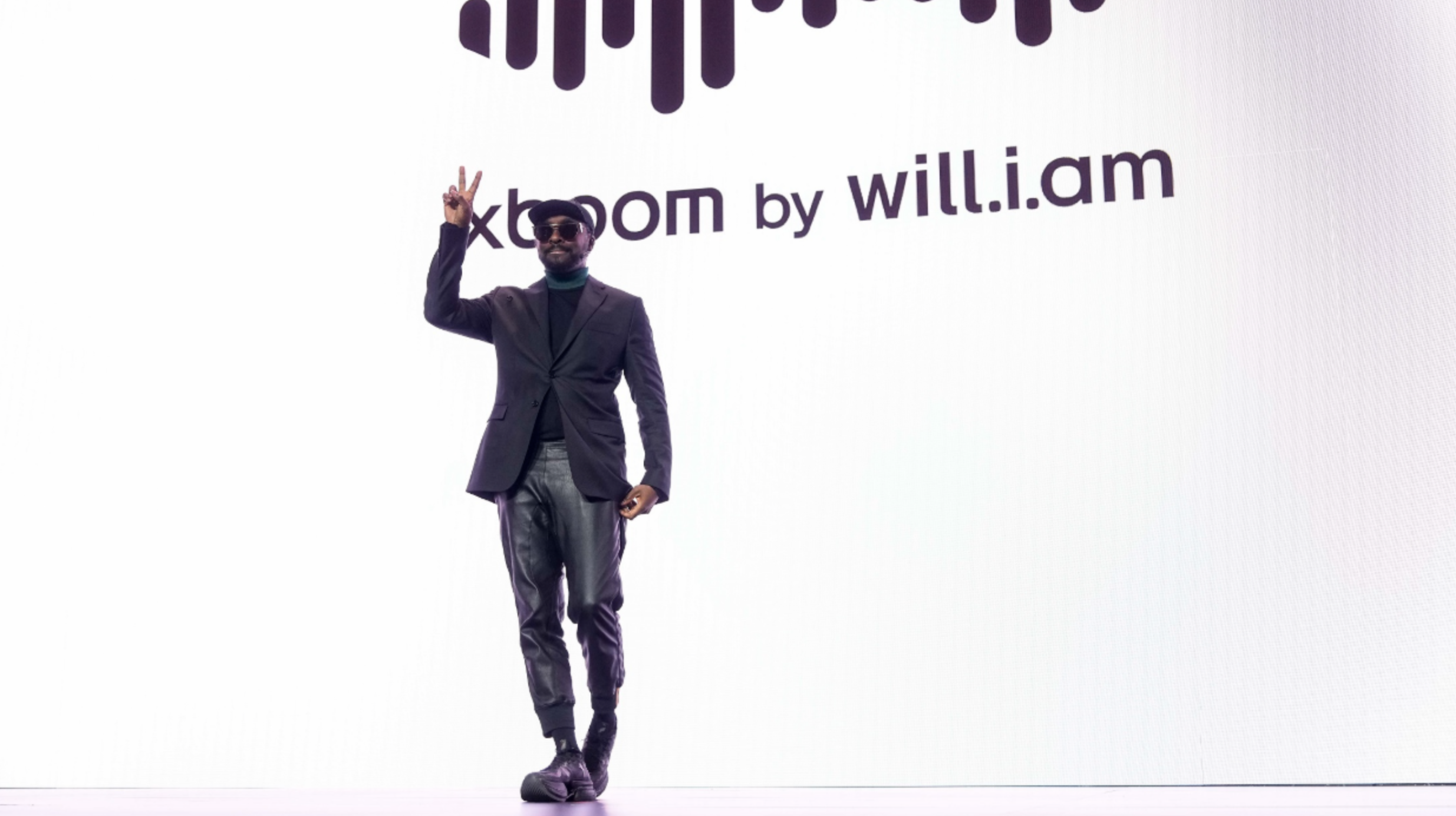 will.i.am wearing all black, making a peace sign, in front of a huge XBoom by will.i.am screen