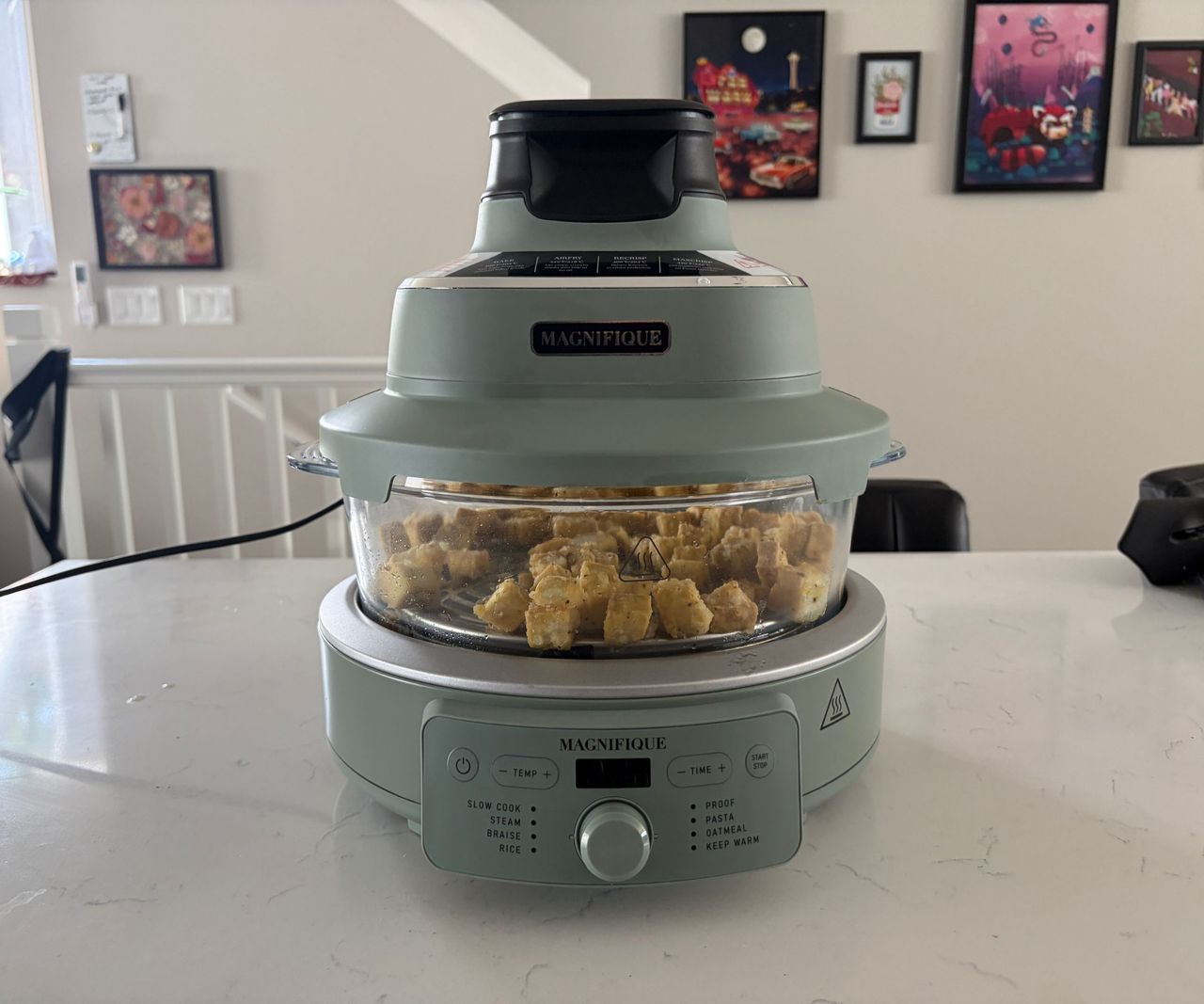 A glass air fryer with a green base on a white countertop filled with tofu.