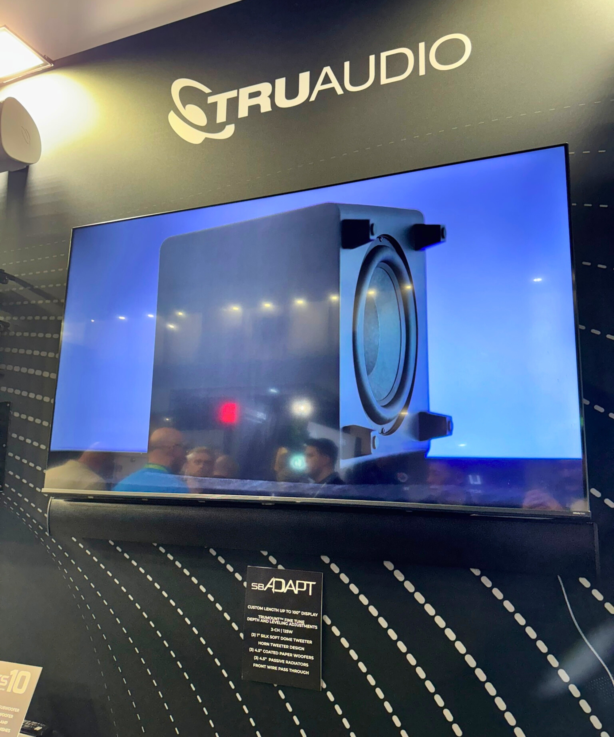 TruAudio display at ISE Barcelona, with custom-fitted soundbar underneath a TV screen displaying a large subwoofer speaker, with an information card on the SB-ADAPT soundbar beneath. Other attendees are visible in the TV screen's reflection.