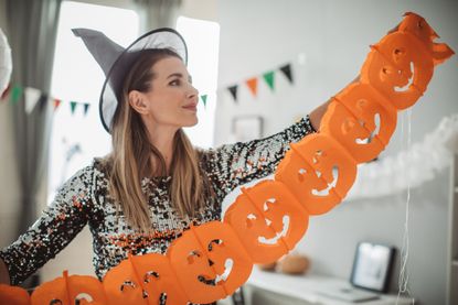 Mature woman in costume decorating home for Halloween, hanging an orange pumpkin garland.