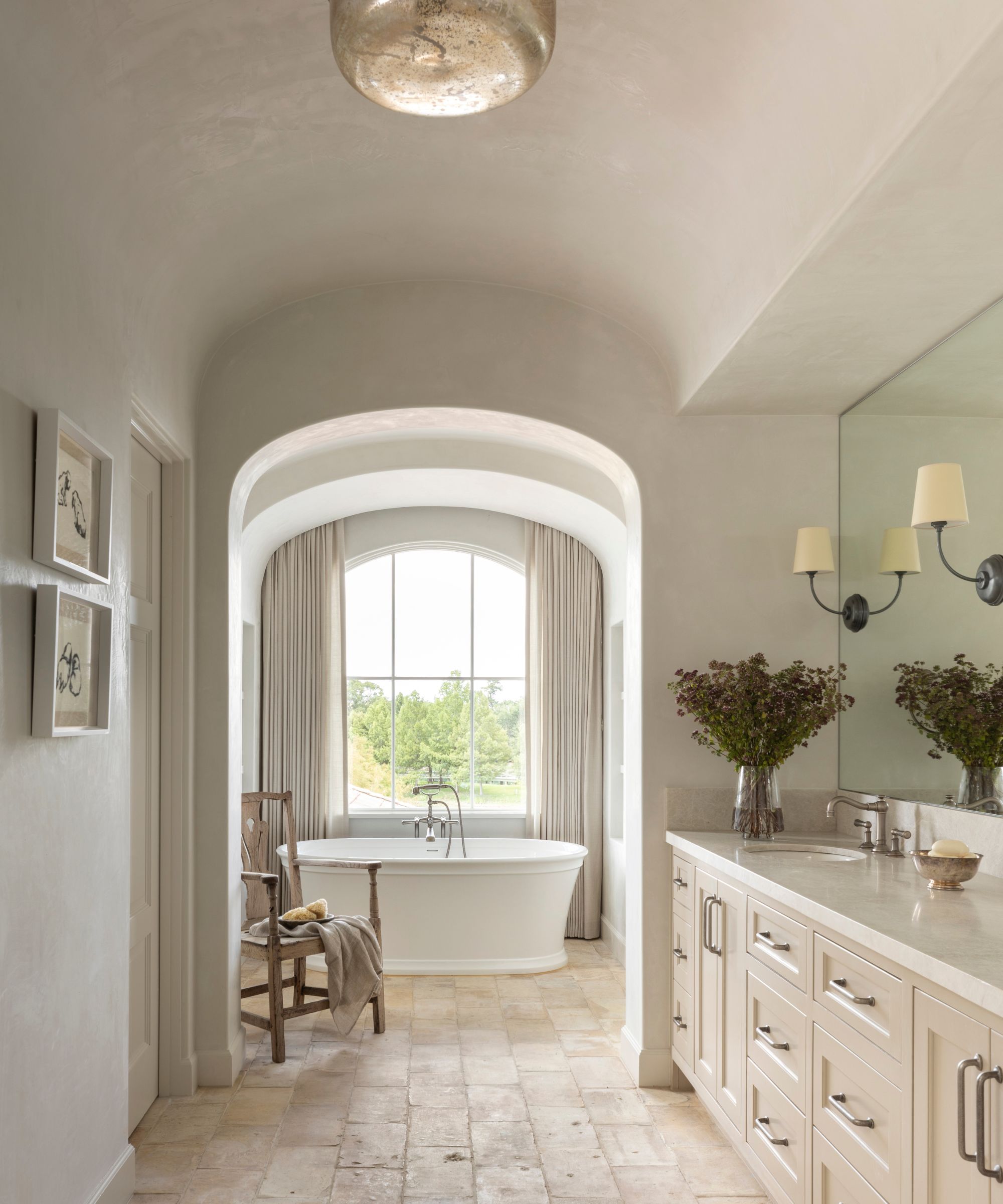 A large bathroom with white plaster walls, an archway leading into a bathtub in front of a large window with neutral curtains, stone flooring, a large vanity and modern rustic decor.