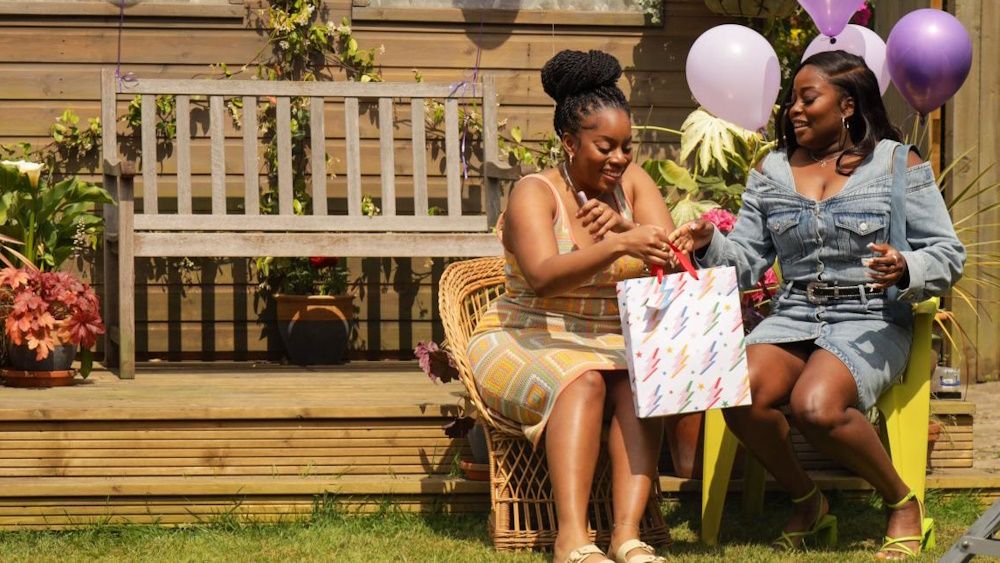 Dionne Brown as Queenie Jenkins and Bellah as Kyazike on a garden bench celebrating a birthday