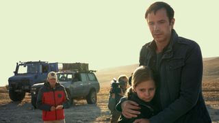 Man hugging a young girl whilst waiting in a desert. In the background there is a large truck, a Jeep, a person in a red jacket wearing a cap and also a man with a big camera.