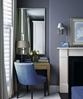 Blue bedroom with dressing table in alcove, mirror on wall, upholstered seat, table lamp and accessories on table, fireplace,