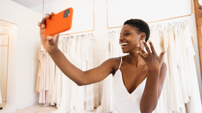 A bride in a wedding dress shop poses for a selfie while holding her hand up to the camera to show her engagement ring