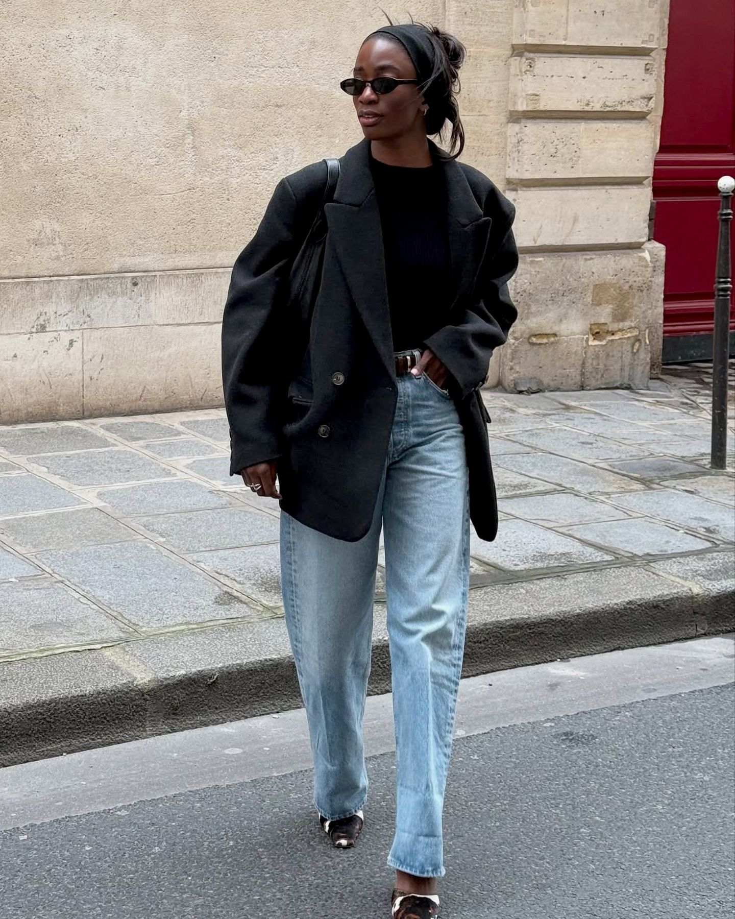 British style influencer Liv Madeline crosses a street in Paris wearing a black headband, black oval sunglasses, an oversize black blazer jacket, a black top, a black belt, light-wash barrel jeans, and cow-print mule heels.