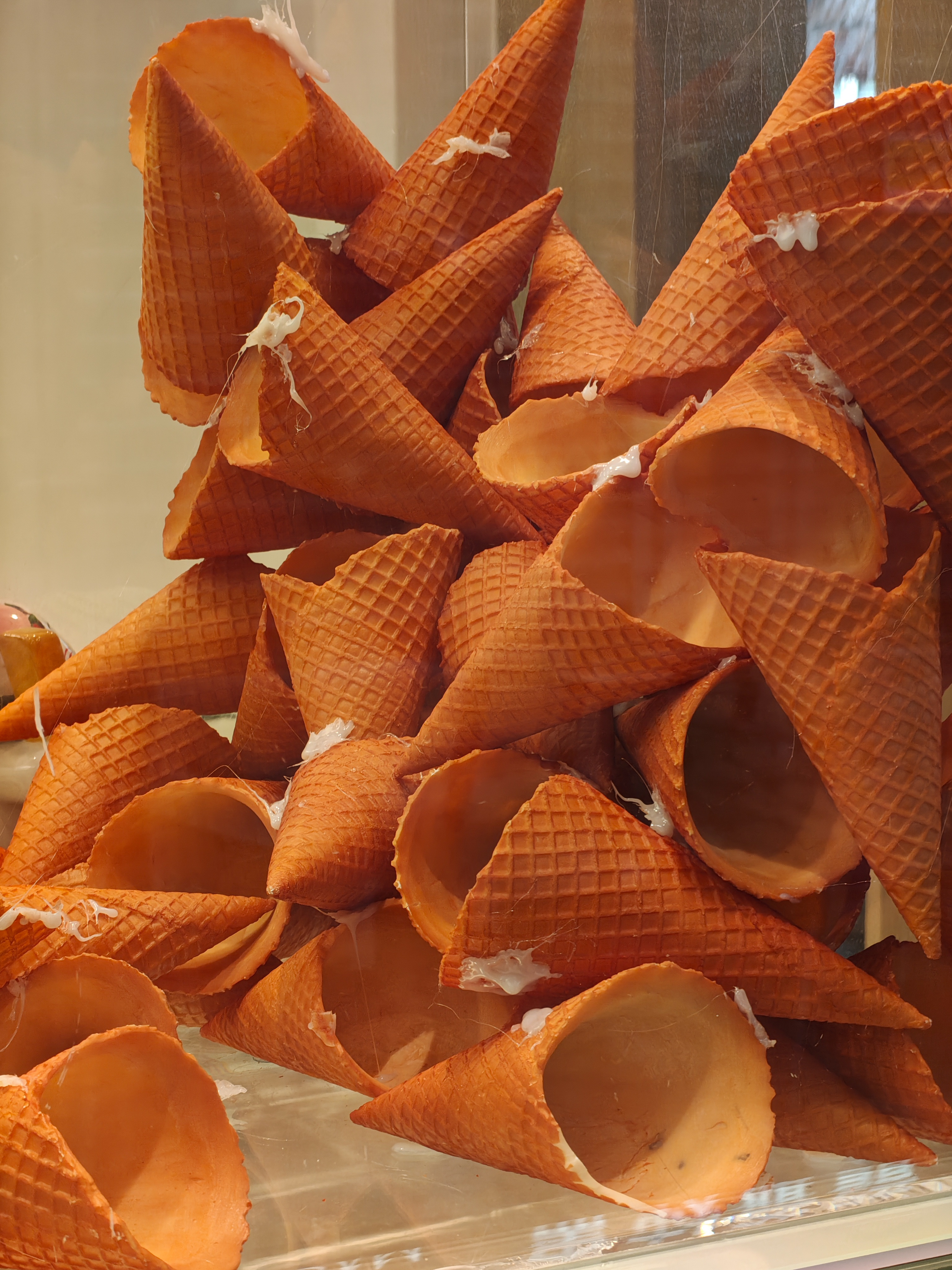 A close-up, high-angle shot of a large pile of orange waffle ice cream cones. The cones are stacked haphazardly, and many have small dollops of white cream or frosting on their edges, making them appear stuck together. The lighting is warm, highlighting the grid texture of the wafers.