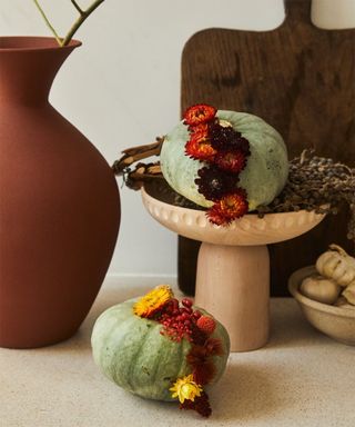 Green japanese pumpkins studded with fresh and dry flowers on wooden bowl with chopping boards in background