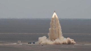 a column of smoke rises up from a flat desert landscape, topped by the spew of fire from a small object flying upward.