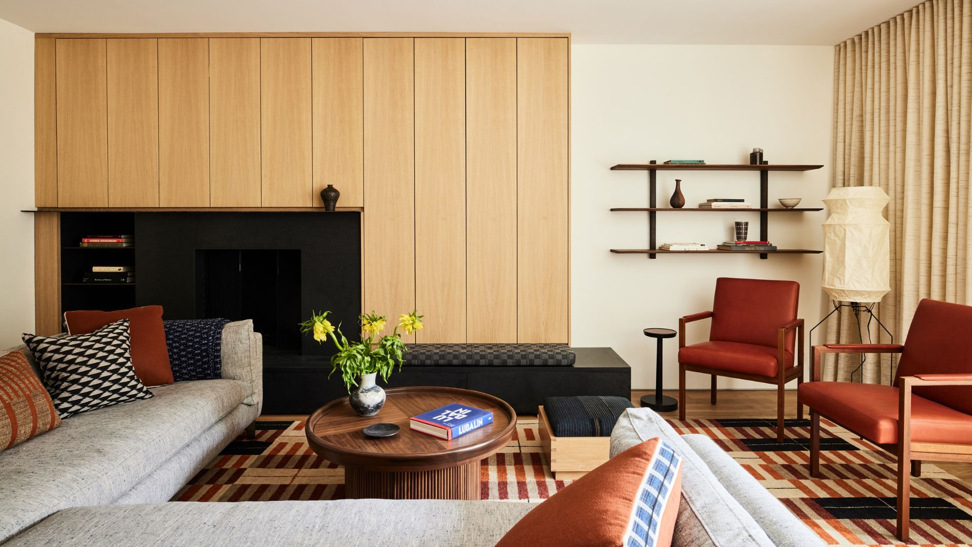 A whit living room with a wood panelled wall, a floating shelf, a floor lamp, a striped area rug, a pair of rust armchairs, a gray sectional, a round wood coffee table, and patterned pillows