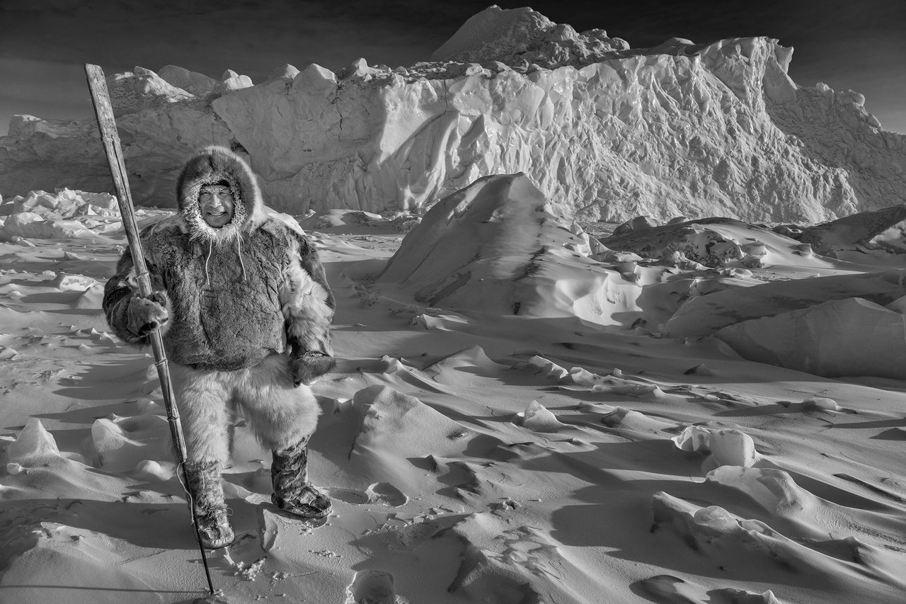 An image of a person in a thick fur outfit standing on a snowy landscape with ice formations. The scene is calm, conveying a sense of solitude