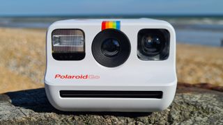 En vit Polaroid Go står placerad på en sten med en strand i bakgrunden.