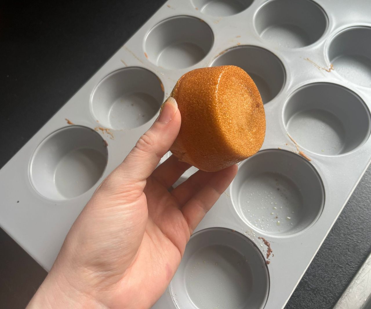 A muffing with a golden base being held above a grey 12-hole muffin tin