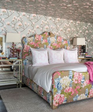 Bedroom with gray and pink blossom wallpaper on walls and ceiling, large green and pink floral bed, mirrored bedside table and matching table lamps, beige patterned carpet