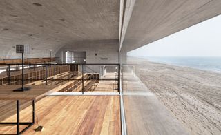 Daytime sea views from the Library large glass front, wooden floors inside the building, black metal table lamp, sand and sea in the distance