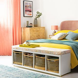 Bedroom with white walls, double bed with yellow and green bedding, stool with storage baskets at the end of the bed