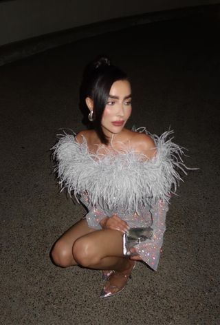 Hanukkah outfit idea is shown in a photo of a woman crouching outside wearing a sparkly blue off-the-shoulder dress with silver heels, silver earrings, and a silver clutch