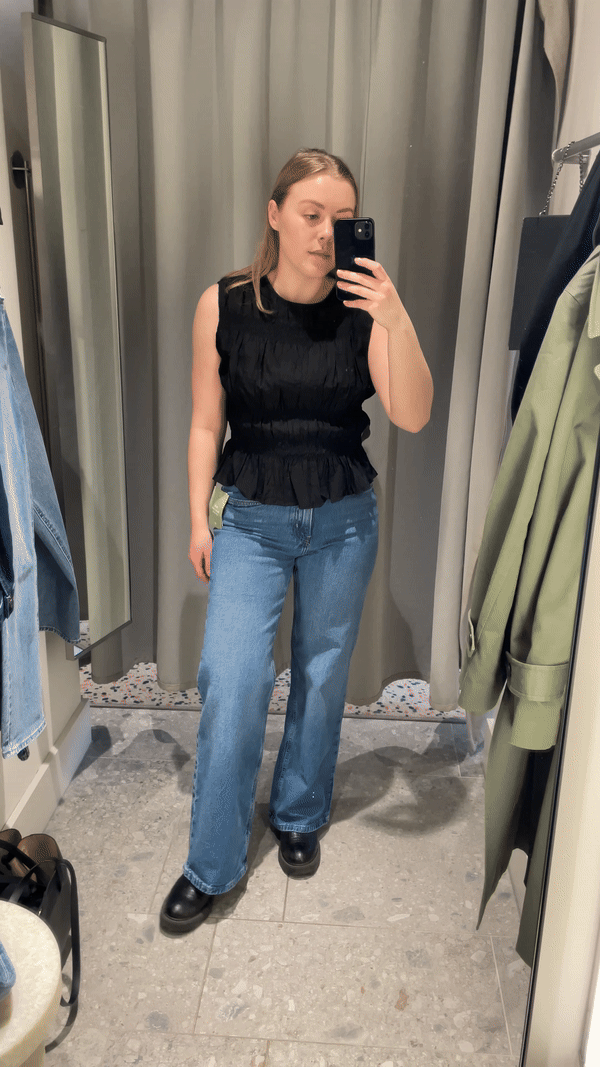 Woman in dressing room wears black top, blue jeans and boots.