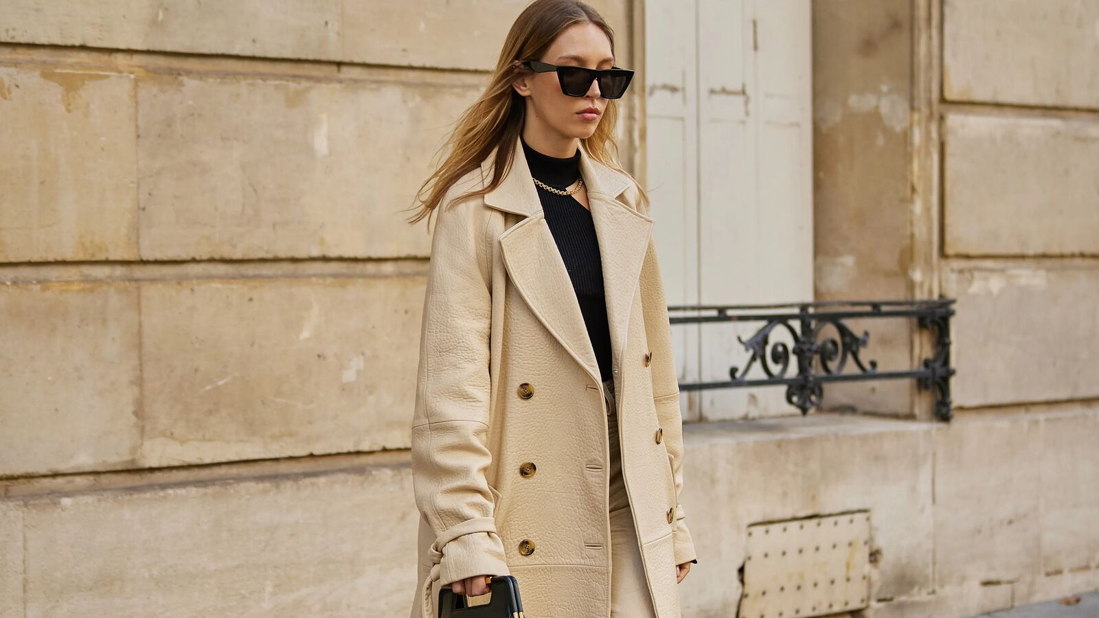 woman wears tan trench coat, tan pants, black sweater, black handbag, and black mule clogs while posing for a street style type of photo