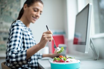 A woman with a long dark pony tail is sat at her work desk. She is eating a take away salad with a folk. There is a computer in front of her.