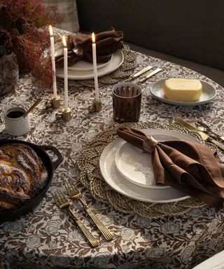 a tonal brown Thanksgiving table setting with a patterned tablecloth, a brown napkin with a gold napkin ring, white tableware, and a rattan placemat