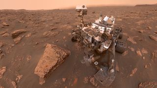Curiosity rover on the rusty red rugged surface of Mars covered with rocks and a hazy red sky. 