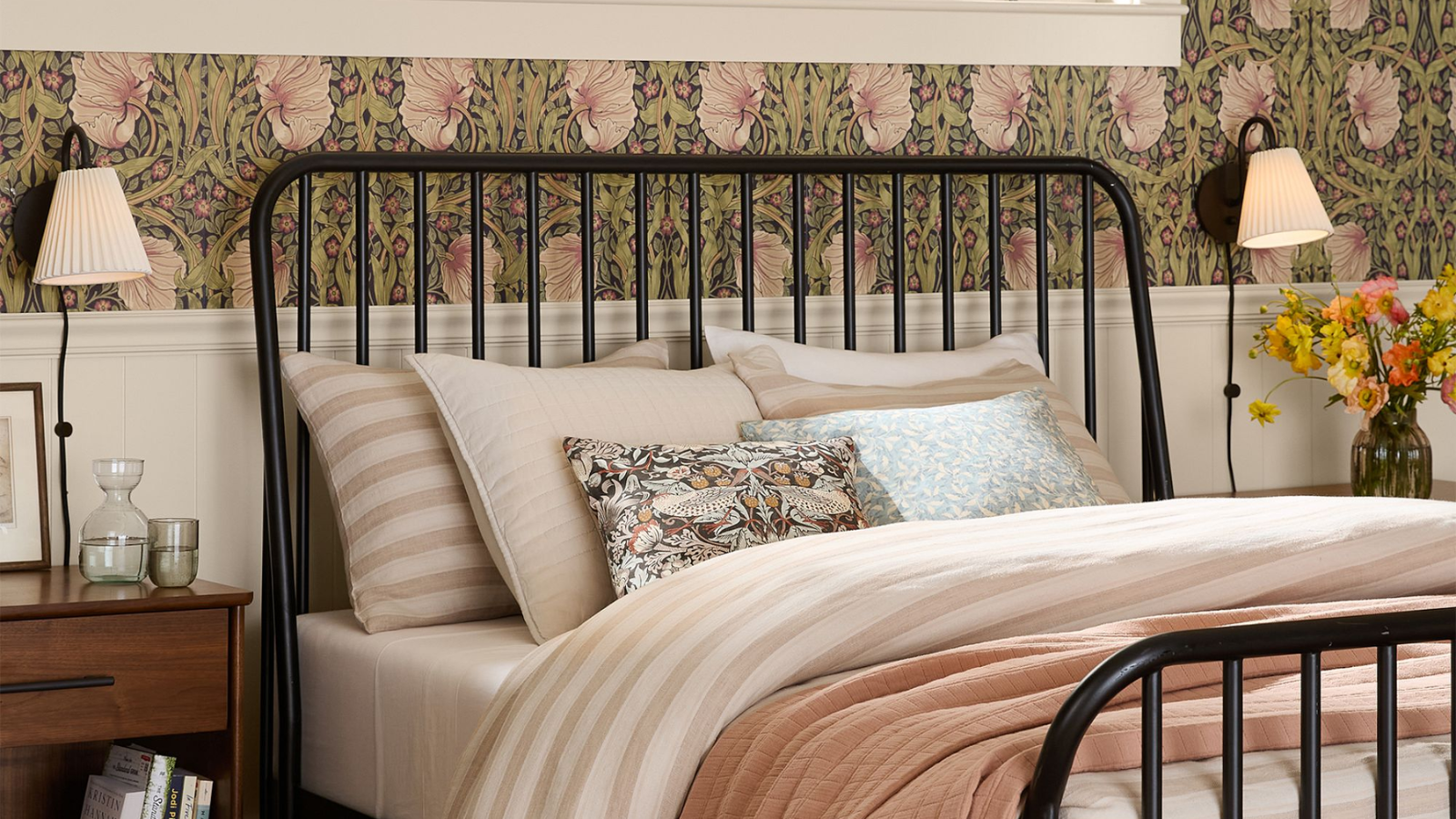 a bedroom with floral wallpaper on the walls, a black metal bedframe, light pink bedding, and two black plug in wall sconces on either side of the bed