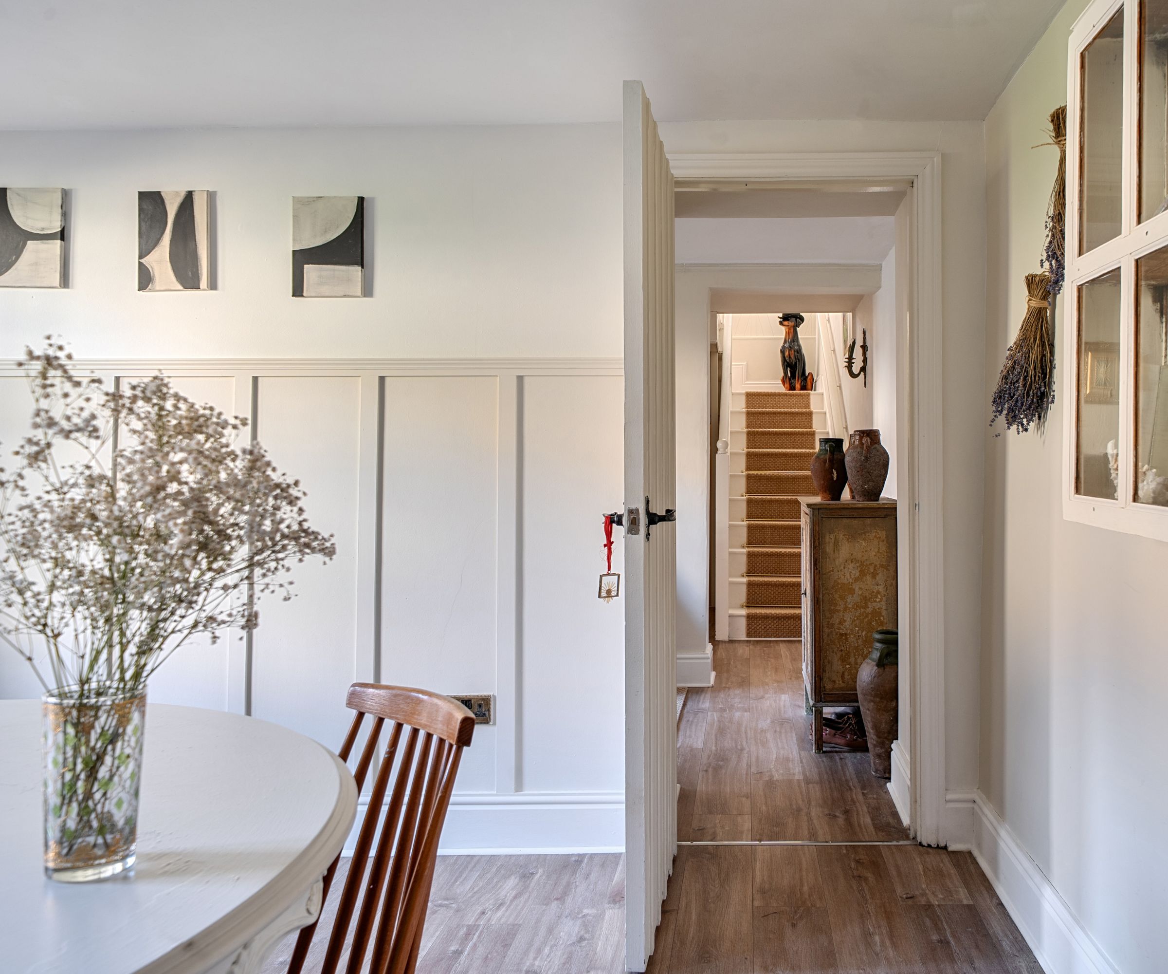 White board-and-batten wall panelling in a bright dining room. Wooden chair, round table, dried flowers, and a hallway with stairs beyond.