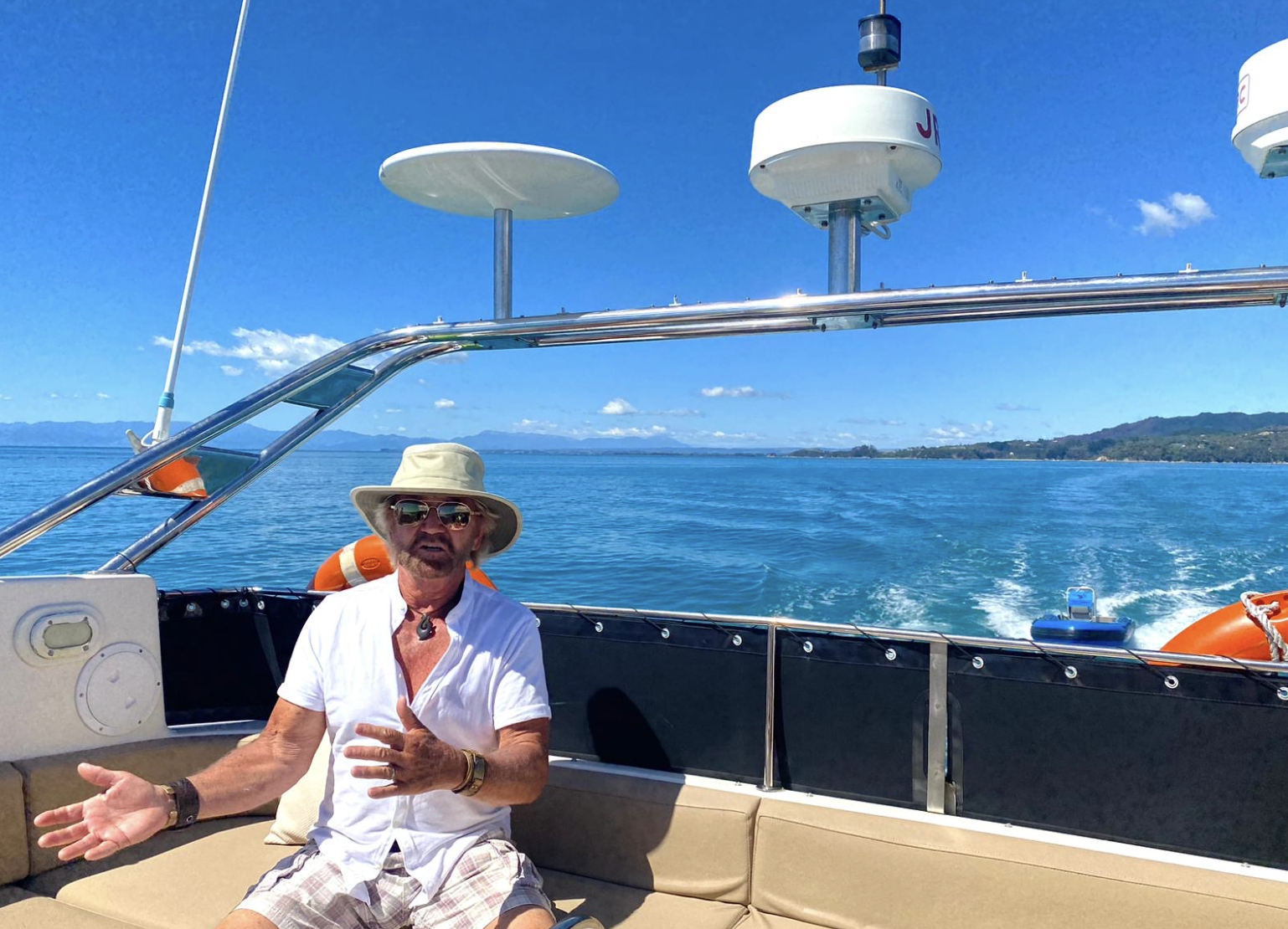 Noel Edmonds sitting on a boat on a sunny day surrounded by clear blue water.