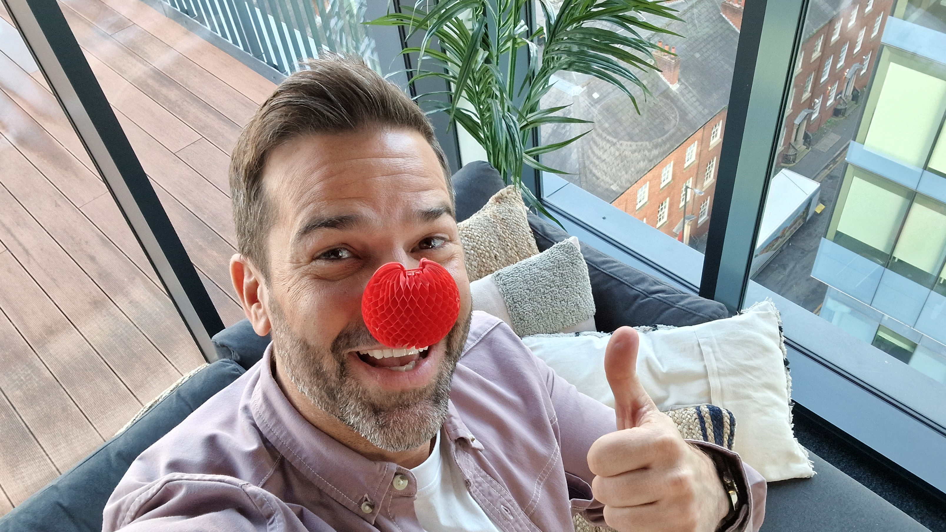 A selfie of Gethin Jones in the Morning LIve studio, sitting on the sofa in front of the window with the street below him, wearing a Red Nose and giving a thumbs-up with his free hand