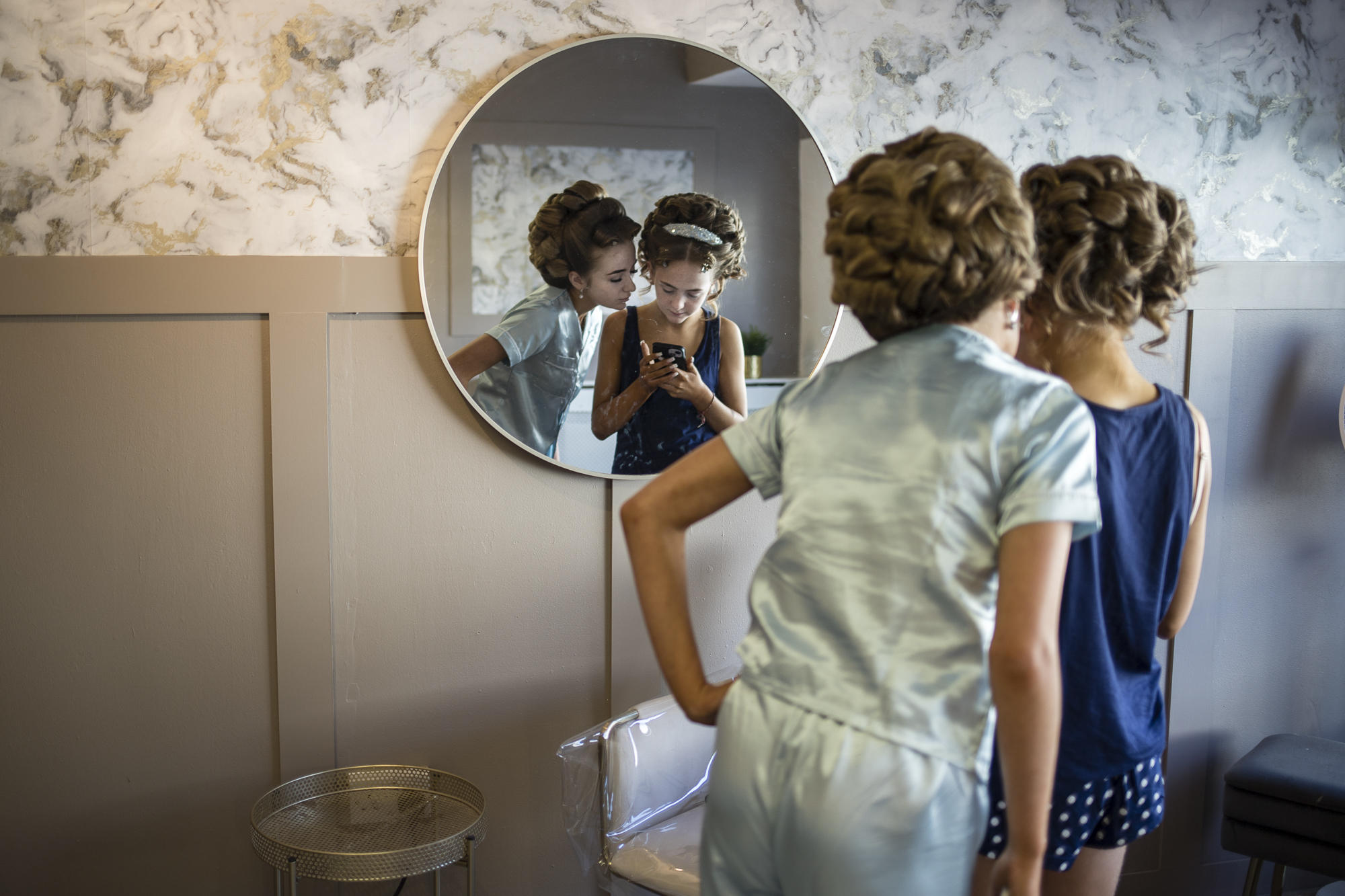 Two young girls with elaborate hair updos stand in front of a round mirror, looking at a mobile phone screen.
