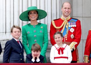 Prince William in a red uniform standing next to Princess kate in a green dress and matching hat on the balcony of Buckingham Palace, with Prince George, Prince Louis and Princess Charlotte standing in front of them smiling