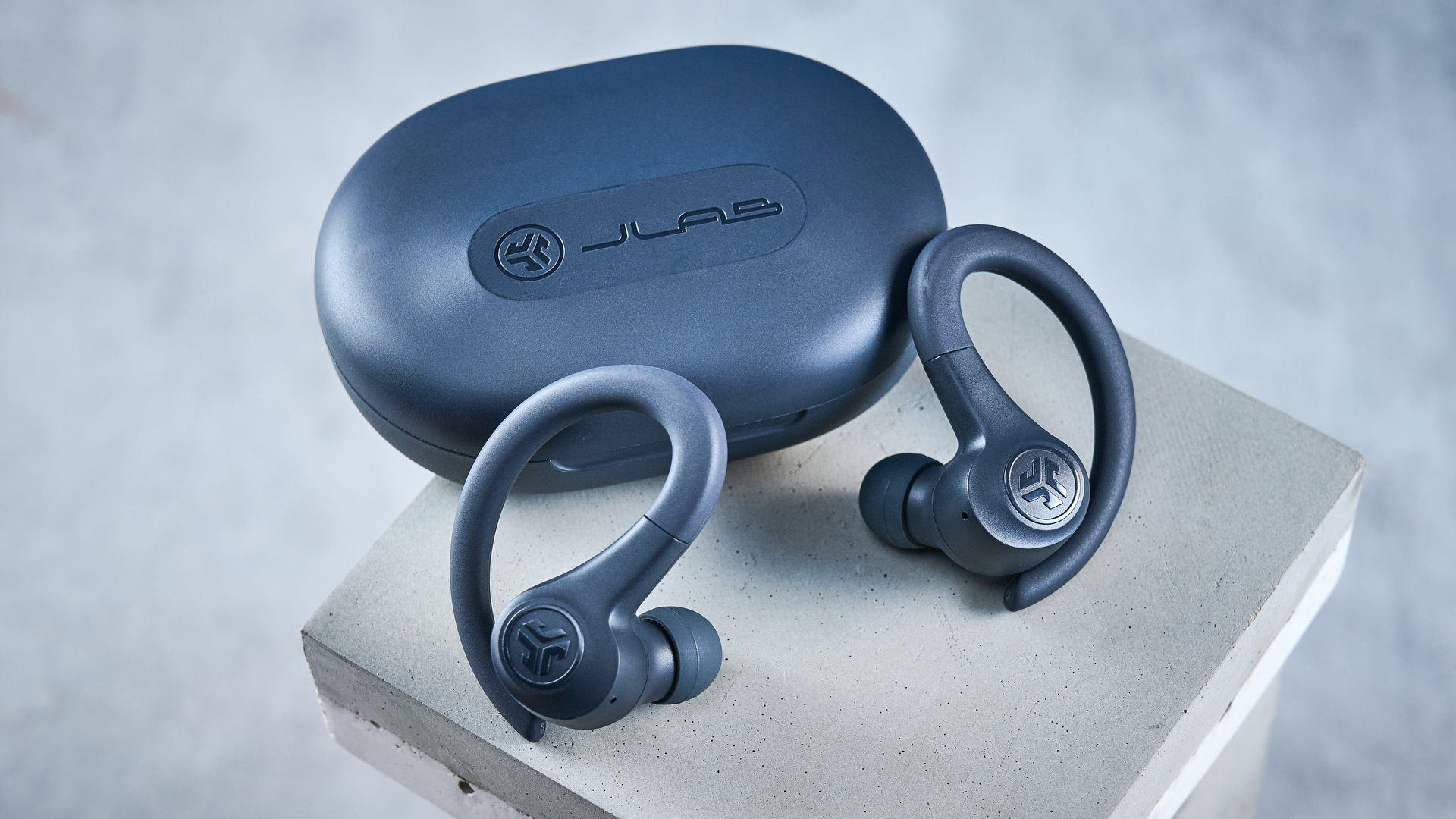 the jlab go sport+ earbuds photographed against the blue tom's guide background, showing charging case and wearing style