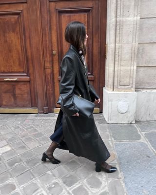 Woman wearing leather trench coat, black shoes, black tights and black leather clutch standing in front of Paris wooden door.