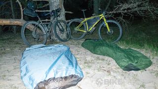 Two bikepacking bikes are propped against trees on a beach while two sleeping bags lie in the sand
