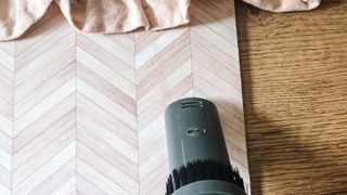 A gray handheld vacuum nozzle in front of a light wooden herringbone placemat on a wooden dining table, with a light pink frilled fabric above it
