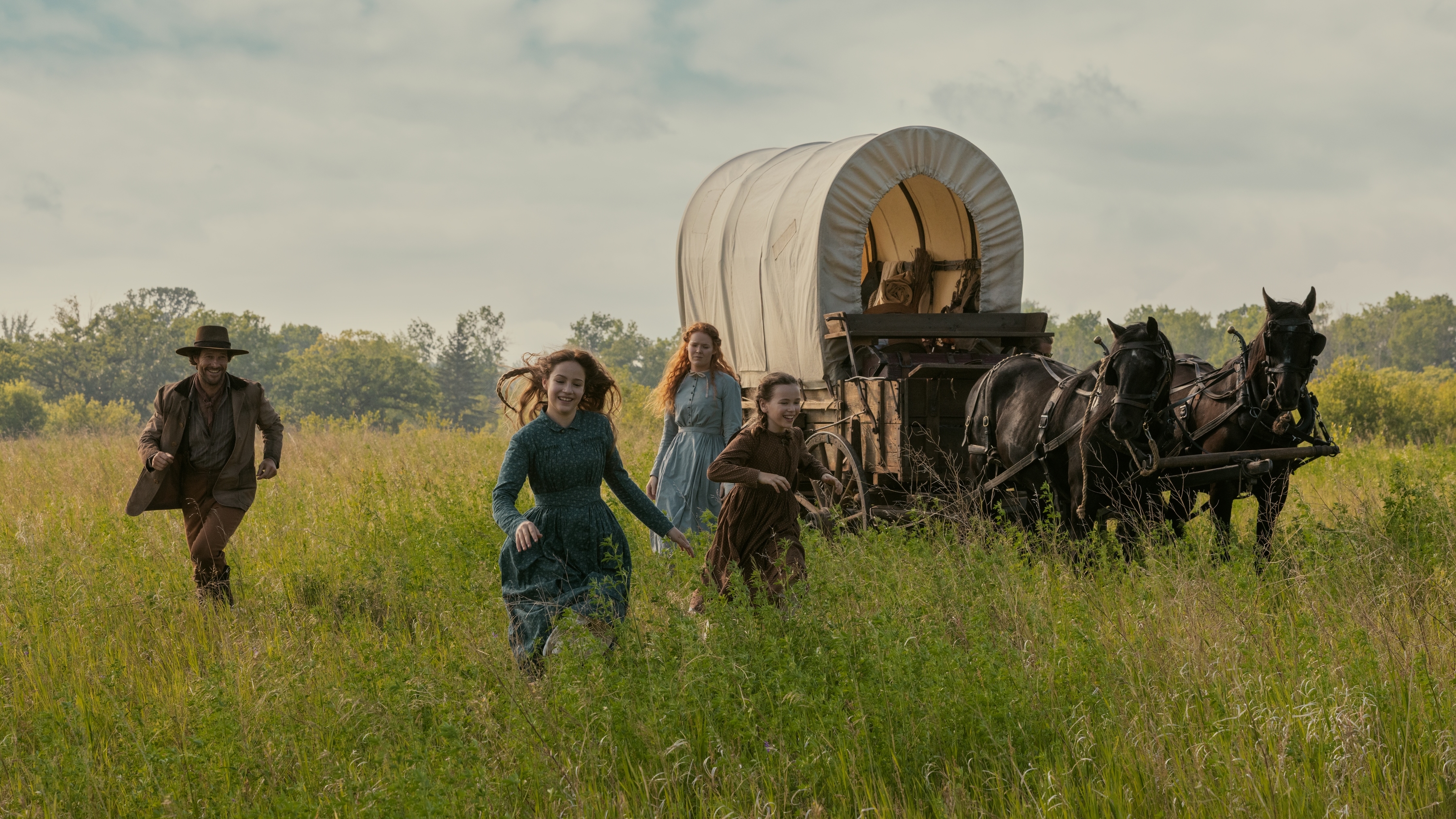 Luke Bracey as Charles Ingalls, Skywalker Hughes as Mary Ingalls, Crosby Fitzgerald as Caroline Ingalls, and Alice Halsey as Laura Ingalls running through a field.