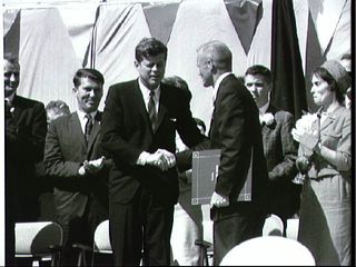 President John F. Kennedy shakes hands with Astronaut John H. Glenn Jr. after presenting him with the NASA Distinguished Service Award. Glenn's wife stands behind him.